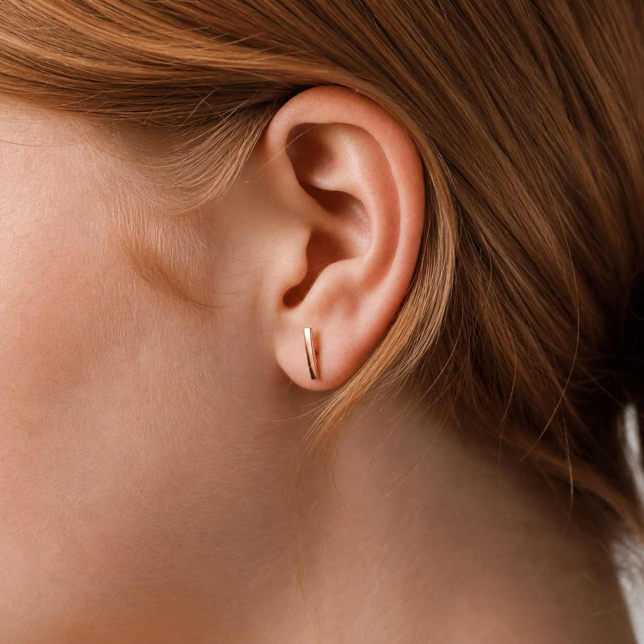 A close-up of a person's ear wearing a slim, vertical rose gold stud earring.